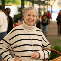 Ann Evans smiling in cream striped turtleneck at outdoor market holding basket of greens (credit Gregory UrquiagaUC Davis)